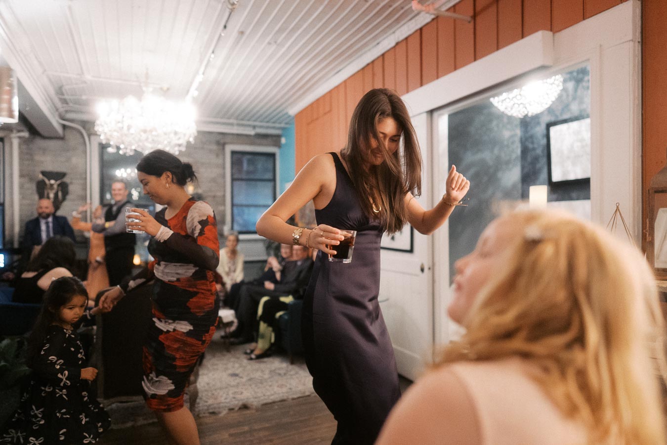 A group of people enjoying a lively dance party in a stylish indoor setting, with a woman in a dark dress dancing in the foreground holding a drink.