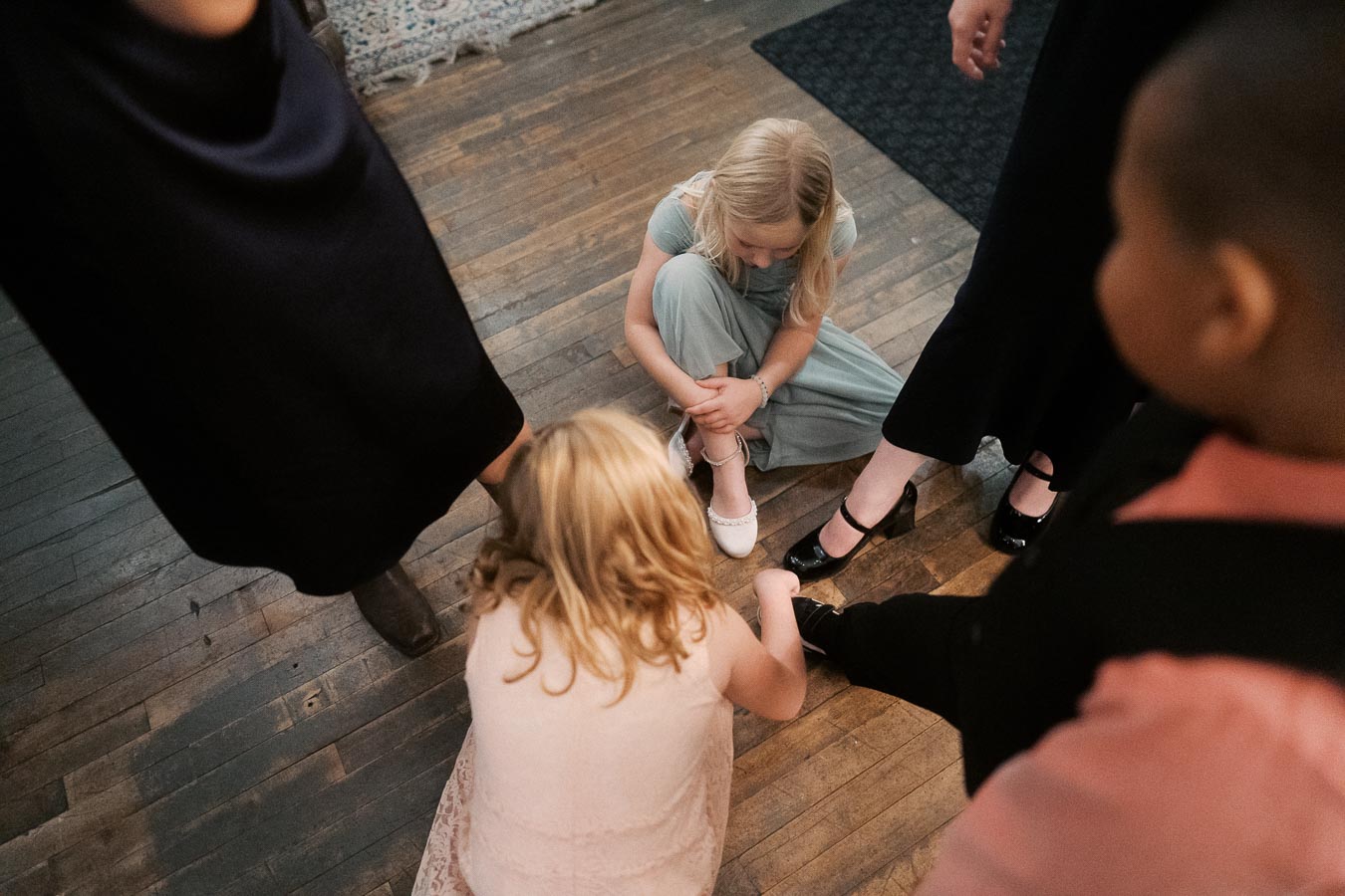 Children playing on a wooden floor surrounded by adults at a social event, with one child sitting and another crouching, creating a lively and engaging scene.