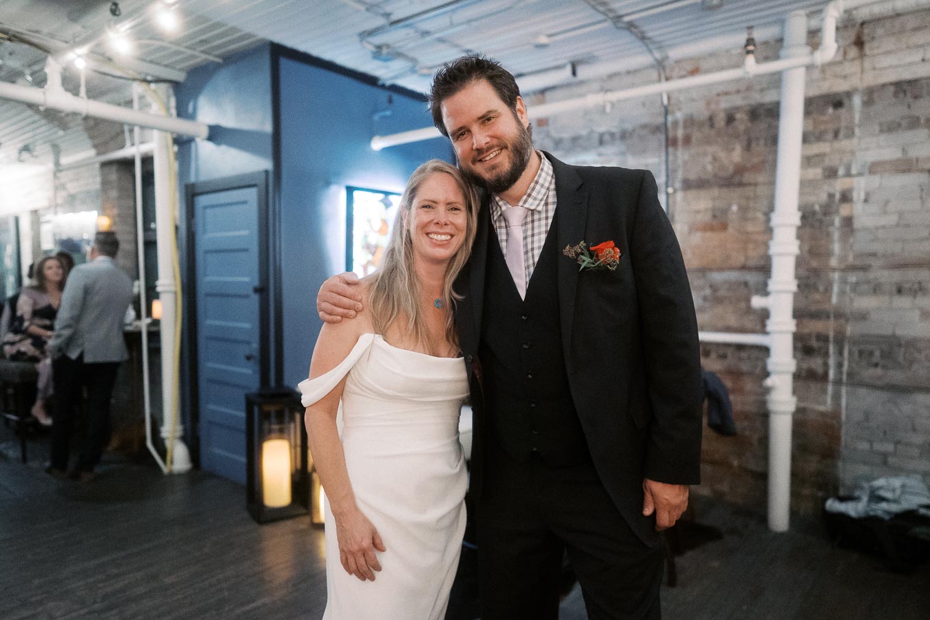 Couple smiling at a wedding reception, with the woman in a white dress and the man in a dark suit, standing in a rustic venue with exposed brick and blue accents.