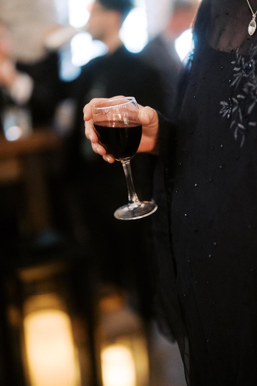 A person holding a glass of red wine at a social gathering, wearing an elegant dark outfit, with blurred background suggesting a warm, festive atmosphere.