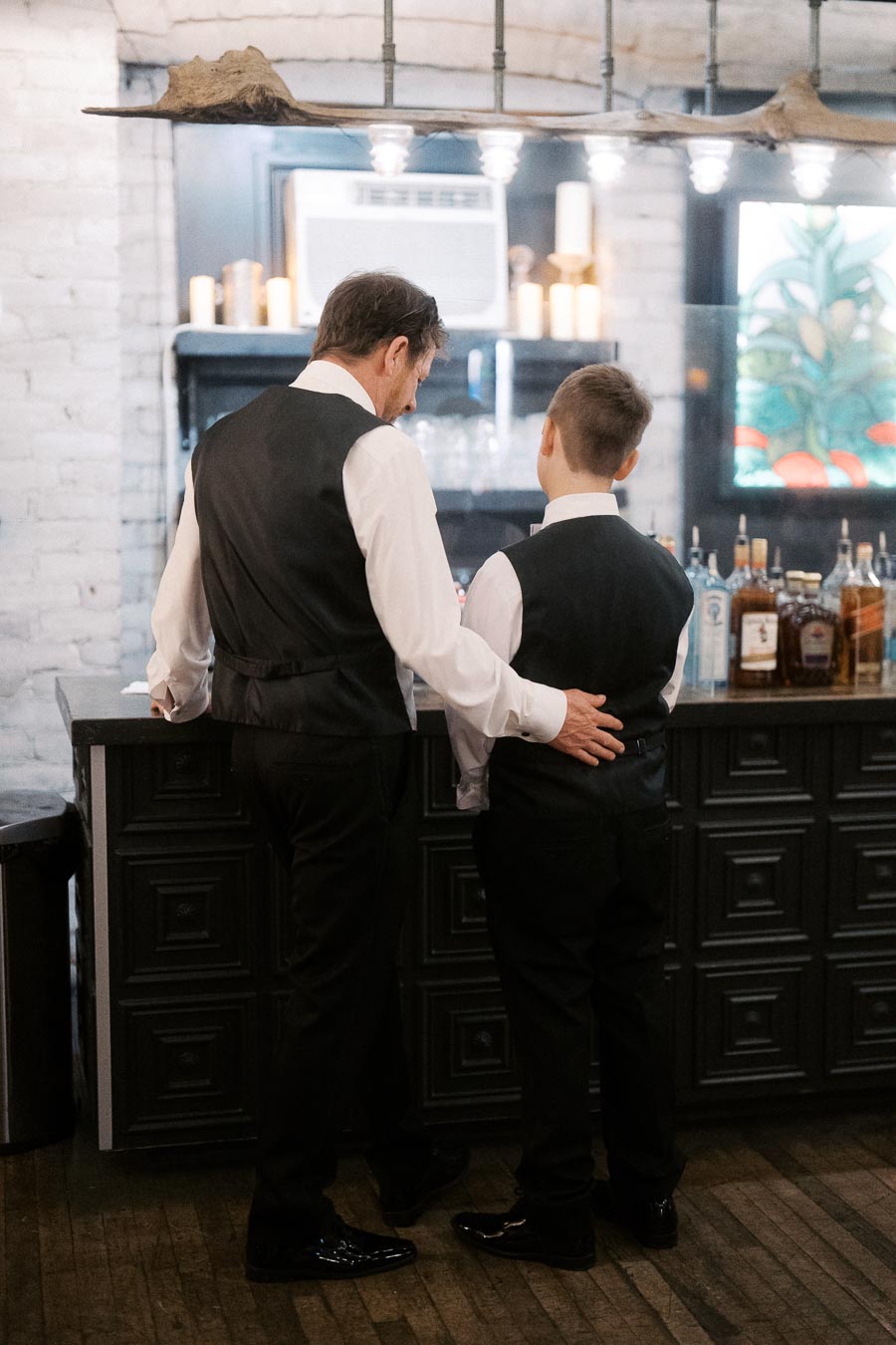 Father and son in matching formal attire standing at a stylish bar, captured from behind.