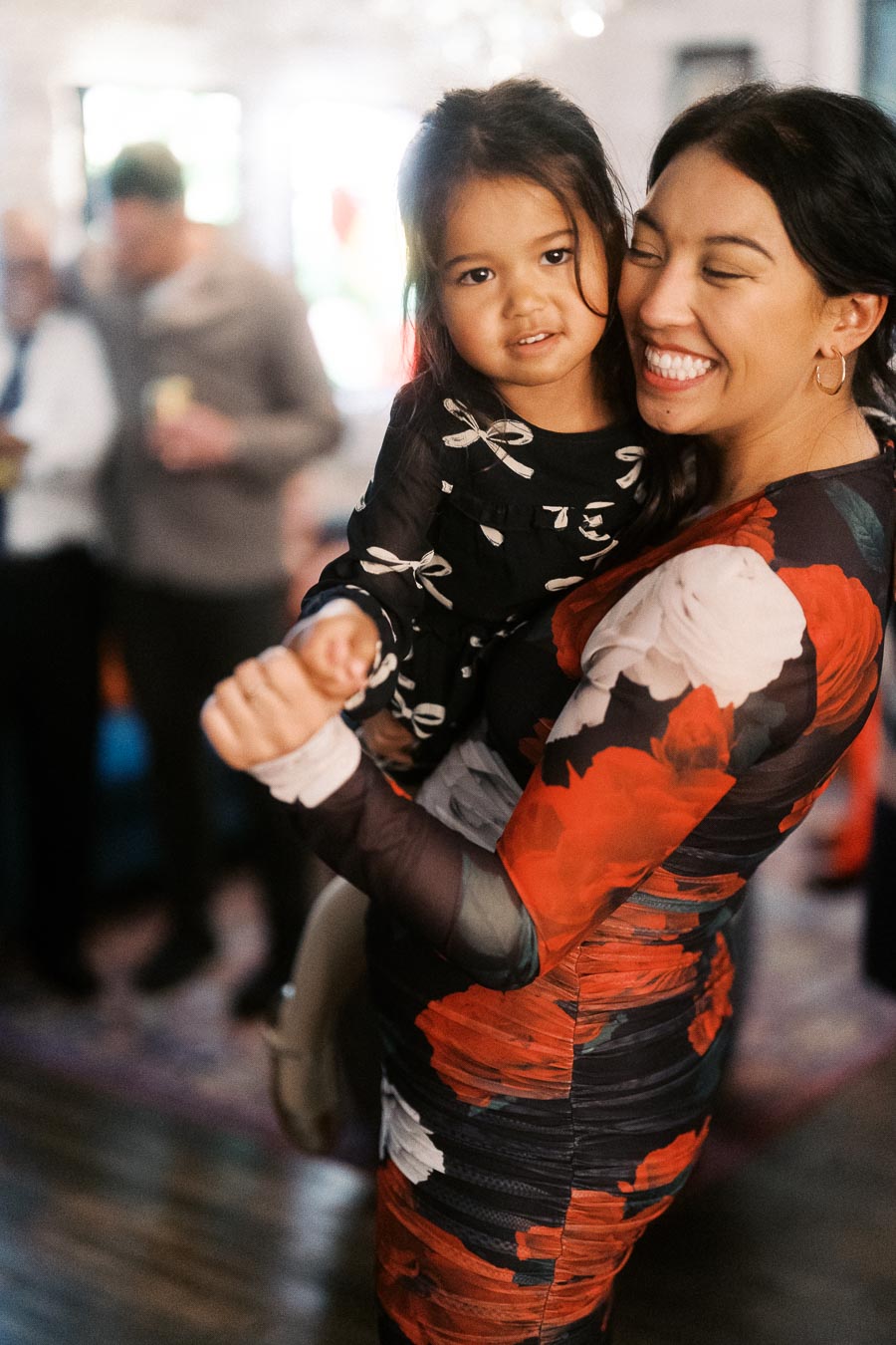 A woman in a colorful floral dress smiling and holding a young girl at a social gathering indoors.