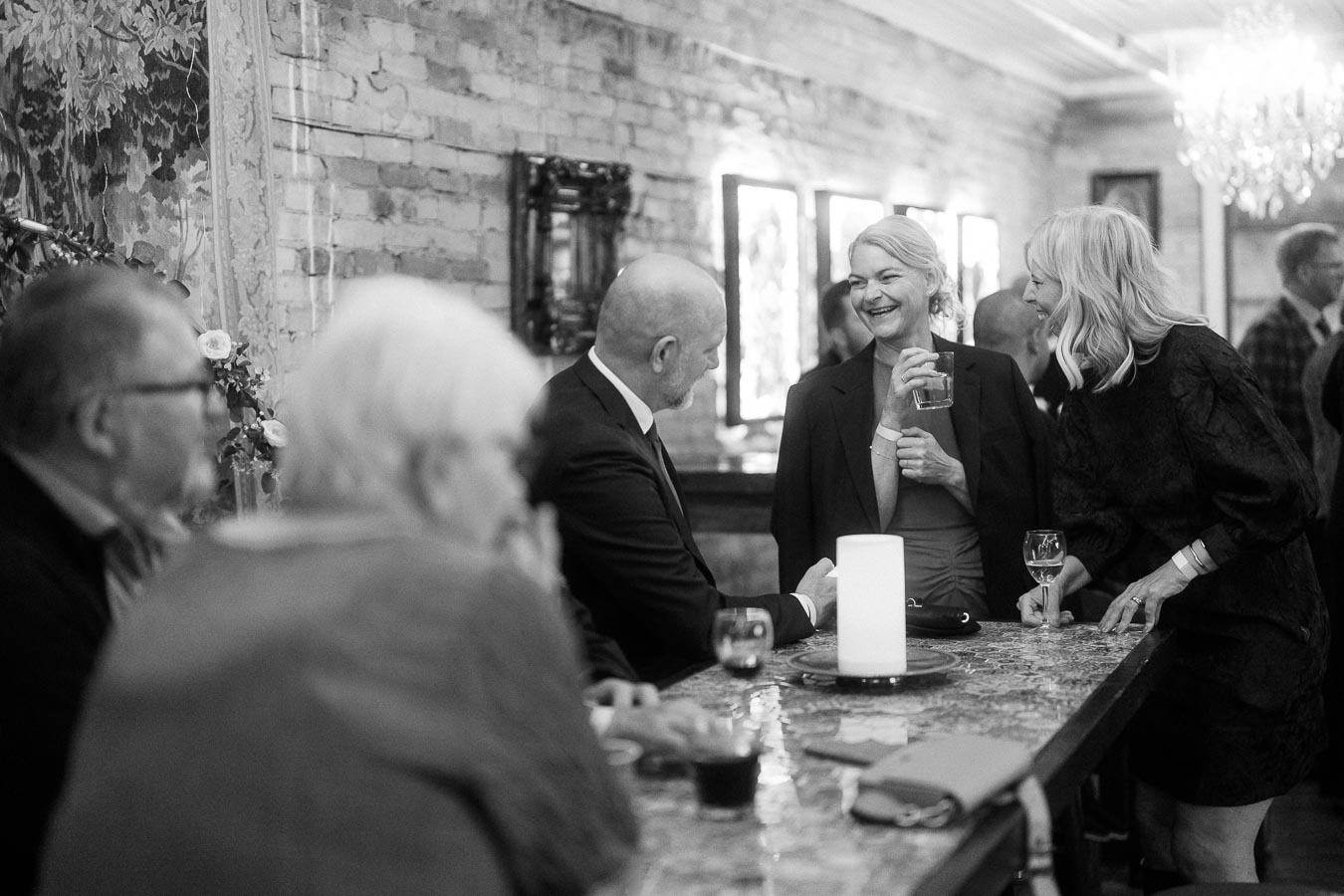 Black and white photo of a lively social gathering at a bar with people in formal attire, engaging in conversation. A woman is holding a drink and smiling warmly at a well-dressed man, while another woman leans in, suggesting a friendly conversation. The setting is elegant, with a rustic brick wall and dim lighting, enhancing the sophisticated ambiance.