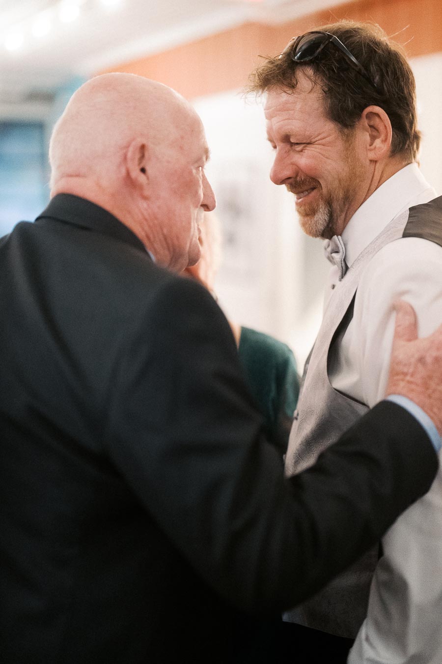 Two men in formal attire share a warm moment at an indoor event, with one smiling as the other gestures towards him, conveying camaraderie and celebration.