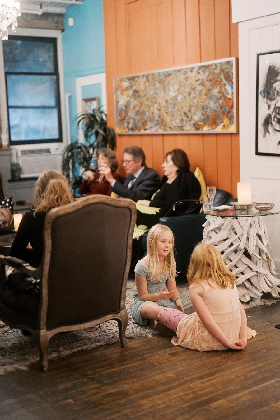 A cozy living room scene with two young girls sitting on the floor engaged in conversation, while adults in the background relax on armchairs. The space is warmly decorated with vibrant art, wooden panels, and ambient lighting, capturing a relaxed, homely atmosphere.