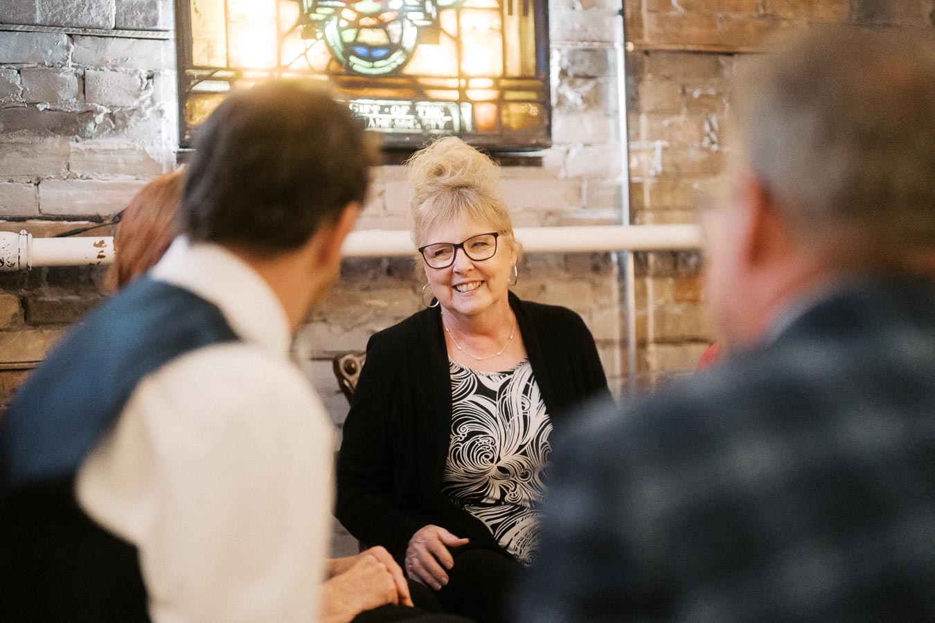 A cheerful woman with glasses and a patterned shirt smiling during a group conversation in a cozy room with brick walls and a stained glass window.