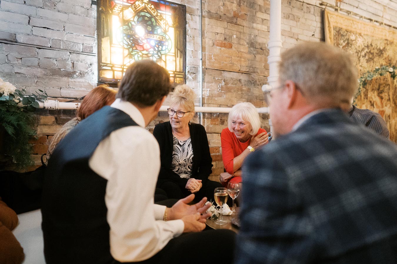 Group of people engaging in lively conversation, sitting together with drinks in hand, inside a cozy room with exposed brick walls and a stained glass window.