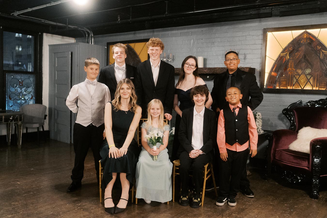 Group of well-dressed people posing together in a formal indoor setting, featuring elegant attire and smiling faces, with decorative elements in the background.