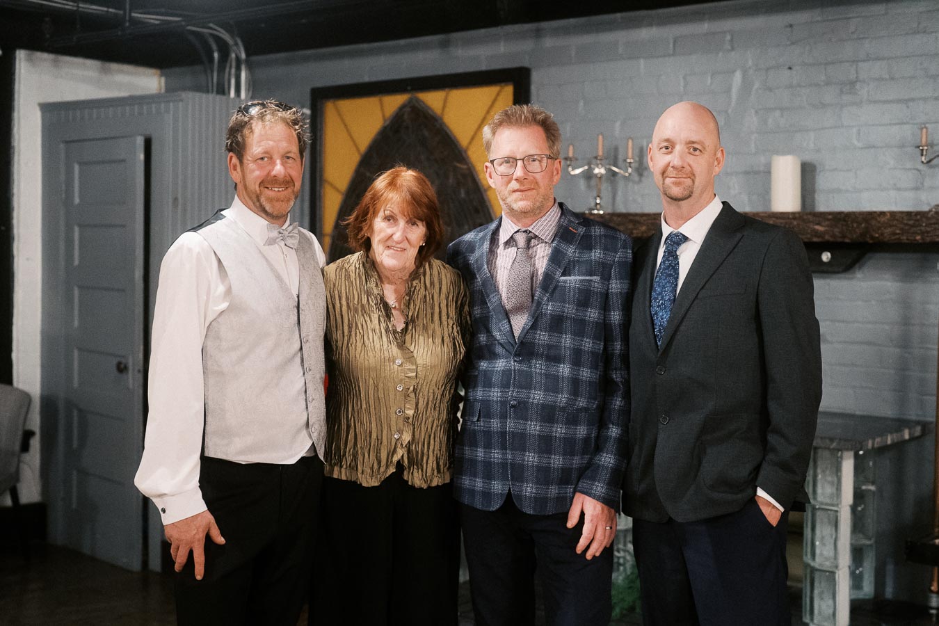 A group of four people dressed formally posing indoors against a gray brick wall with a decorative background, conveying a sense of celebration or gathering.