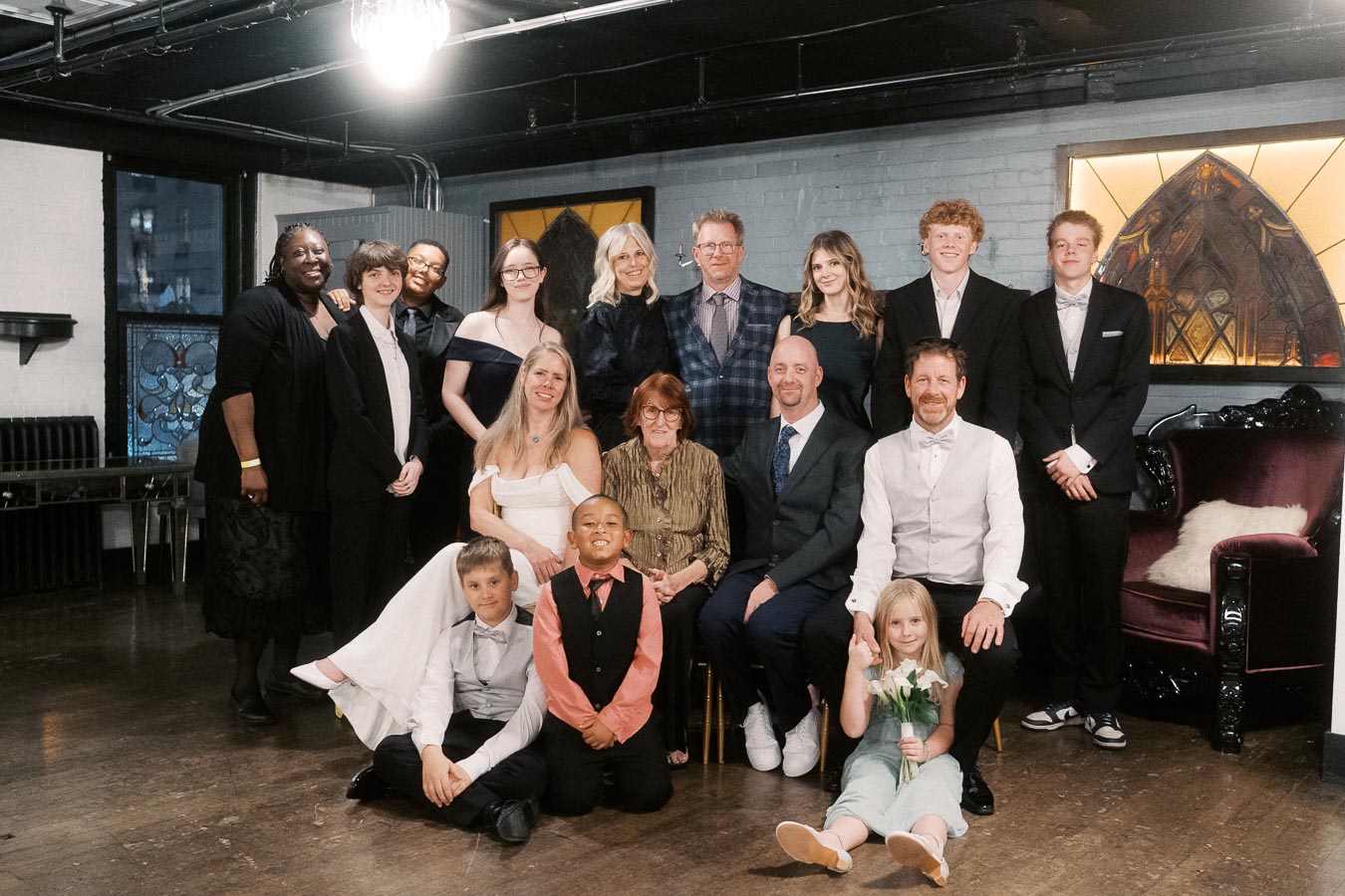 A diverse group of well-dressed people posing together in a modern room with stained glass windows, featuring a mix of adults and children, dressed formally for a special occasion.