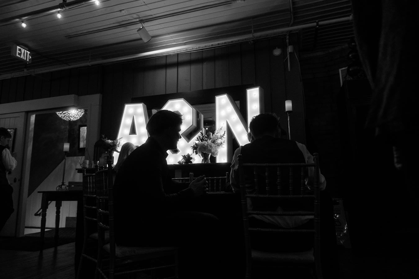Silhouetted people at a dimly lit event, sitting near illuminated marquee letters A&N with floral arrangements, indoor setting with exposed beams and ambient lighting.