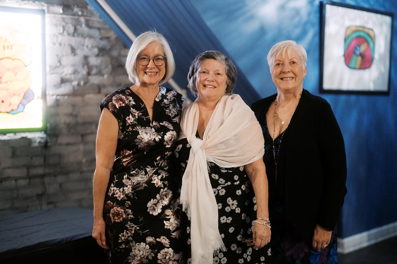 Three smiling women posing together at an event, standing against a blue wall with artwork displayed in the background. The women are dressed in floral and black attire, conveying a joyful and elegant atmosphere.