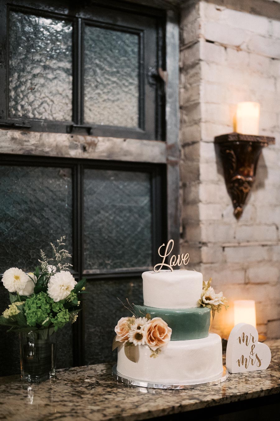 Elegant three-tiered wedding cake with floral decorations and Love topper, set on rustic counter next to a vintage window and lit candle ambiance.