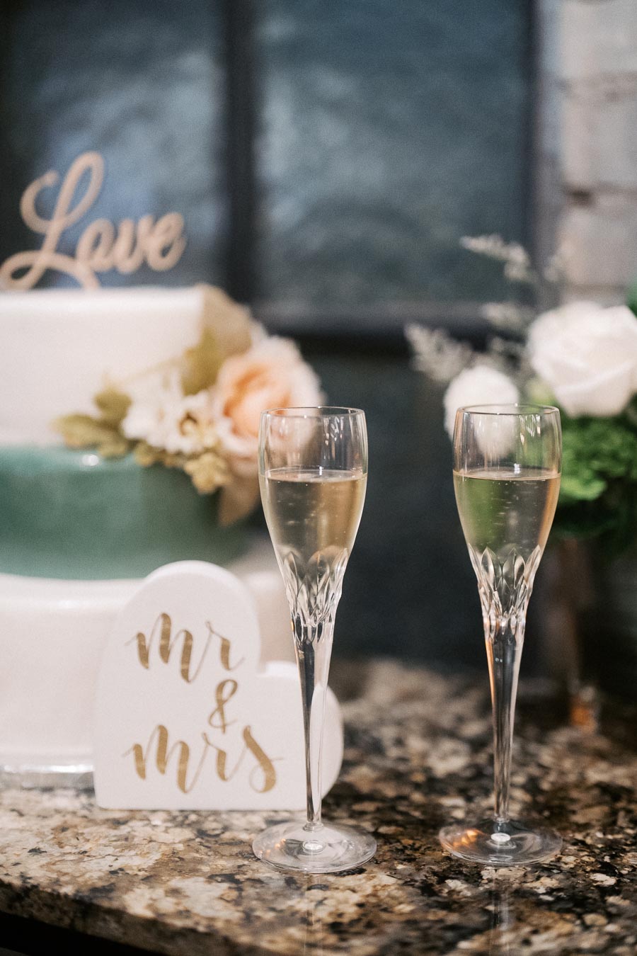 Elegant wedding setup featuring a two-tiered cake decorated with flowers and a Love topper, accompanied by two crystal champagne flutes and a heart-shaped Mr & Mrs sign on a granite countertop.