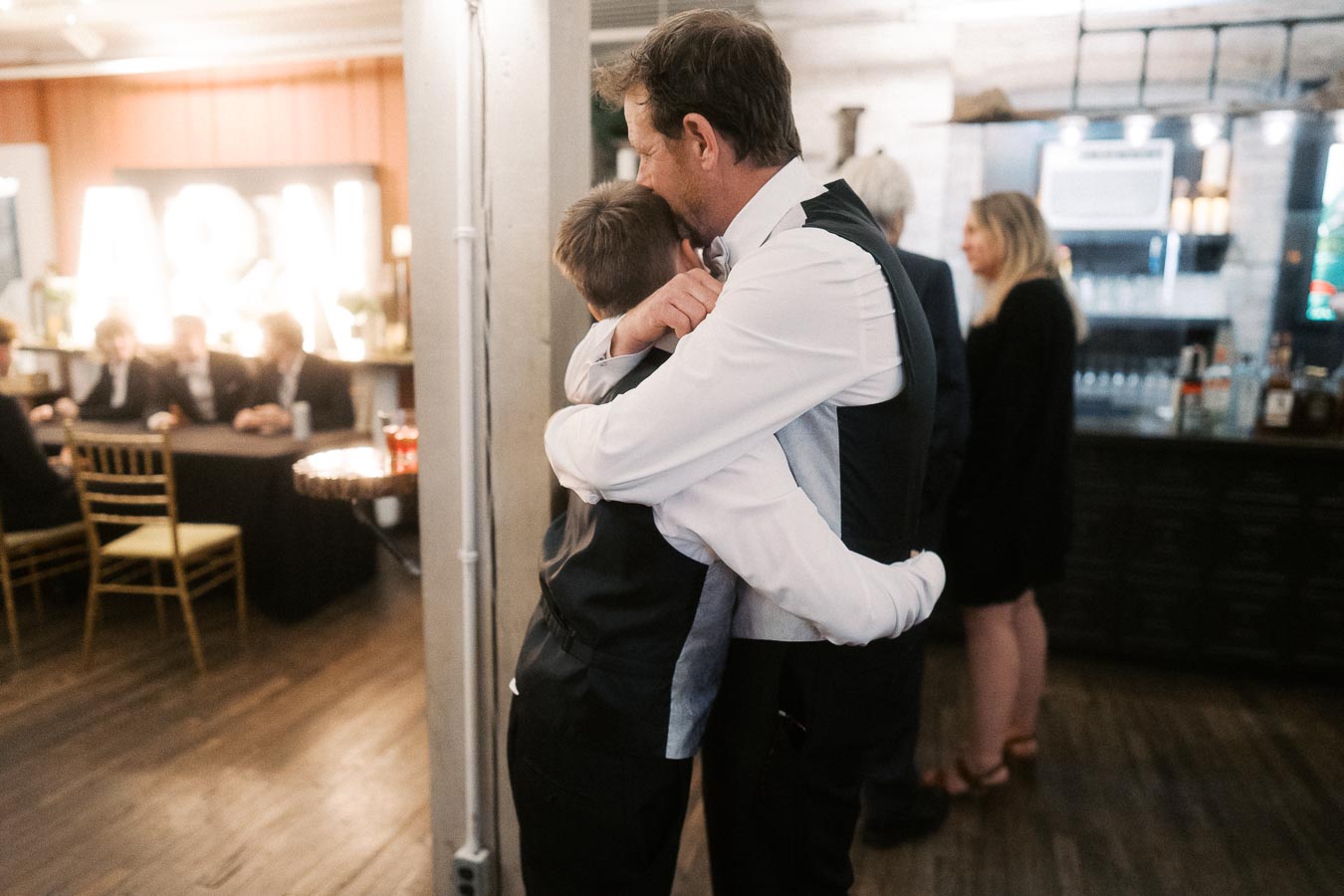 Father and child embracing in a warmly lit event venue, with dressed-up attendees conversing in the background.