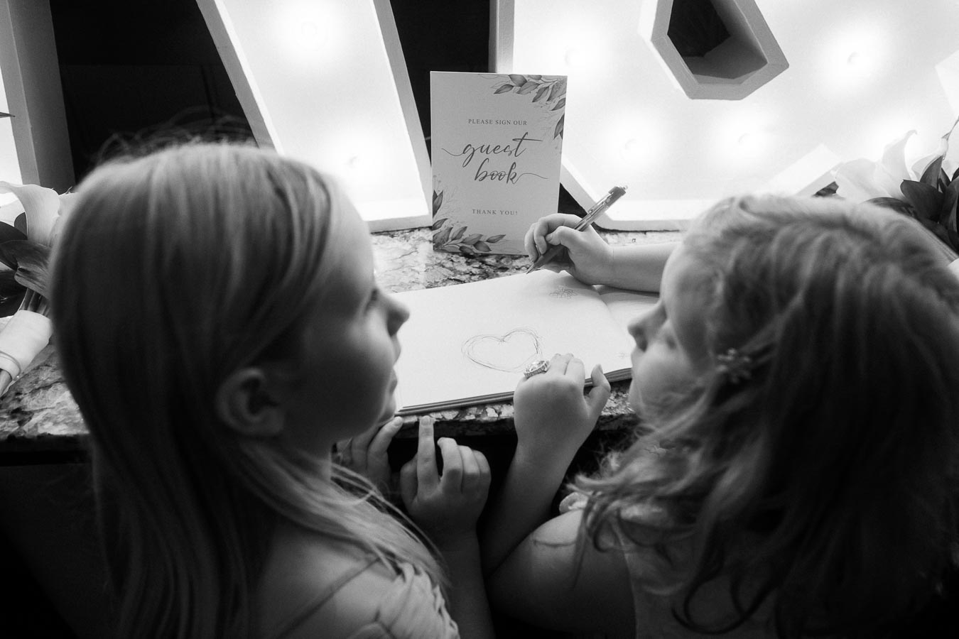 Black and white photo of two young girls standing at a wedding guest book table. One girl is drawing a heart on the open guest book page with a pen. A decorative Please sign our guest book sign is displayed in the background, illuminated by soft marquee lighting.