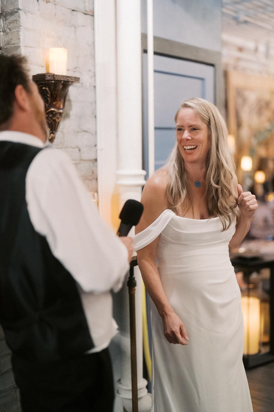 Smiling woman in a white dress enjoying a lively conversation with a man holding a microphone at an indoor event.