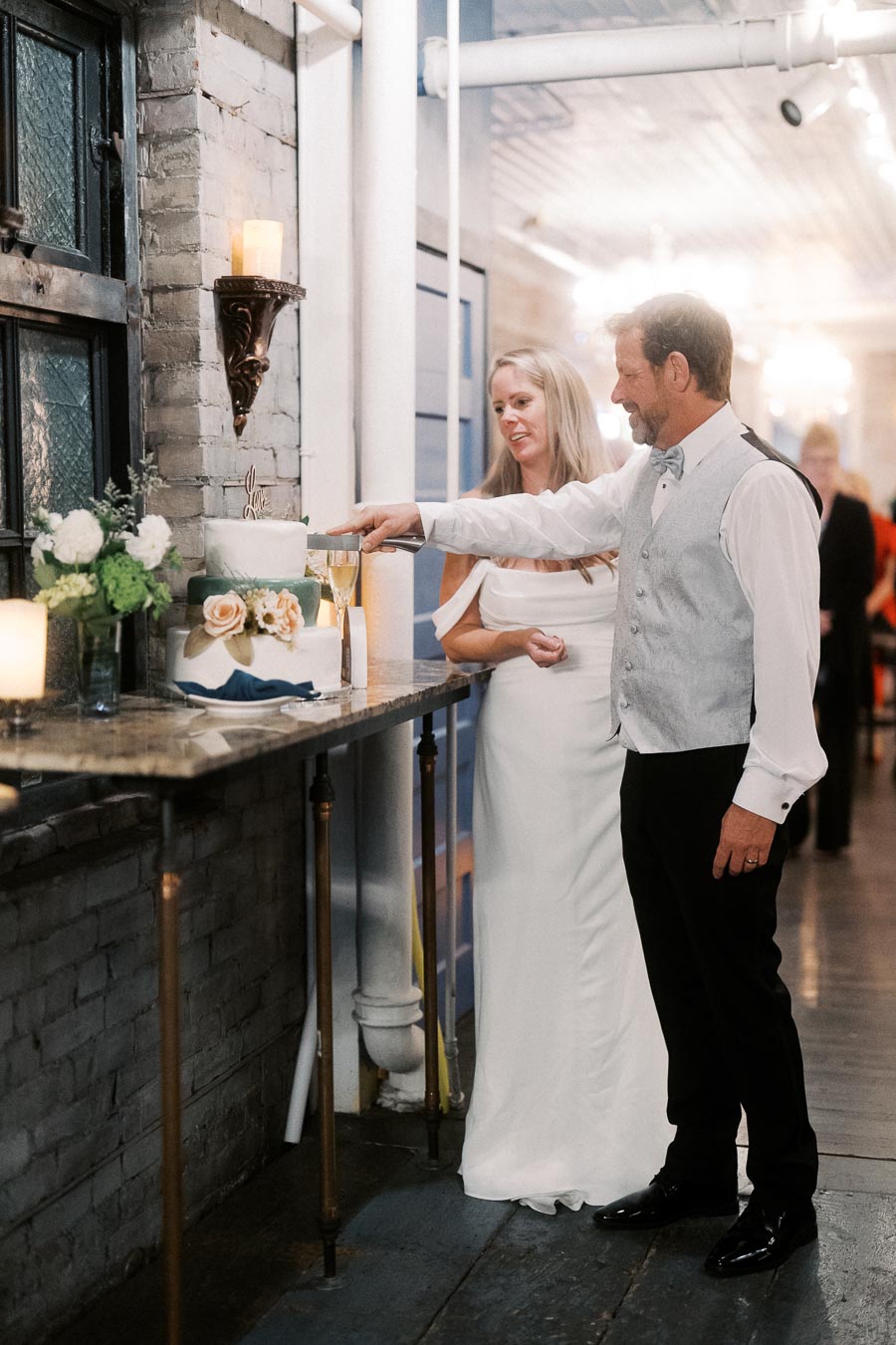 Bride and groom cutting a wedding cake in an elegant venue, surrounded by candles, flowers, and soft lighting.