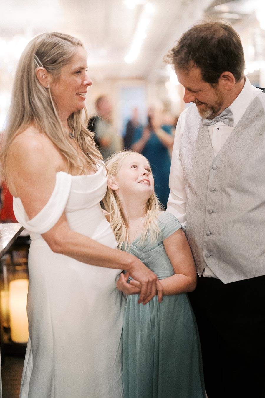 Elegant wedding ceremony with a bride in a white gown, a groom in a vest and bow tie, and a young girl in a light blue dress sharing a heartfelt moment.