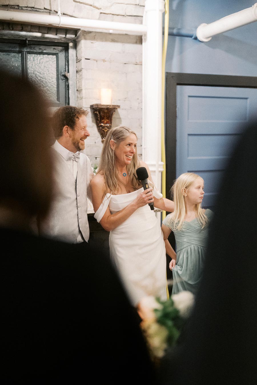 A joyful couple in formal attire, with a woman holding a microphone, stands beside a young girl at a celebratory event in a warmly lit room with exposed brick walls.