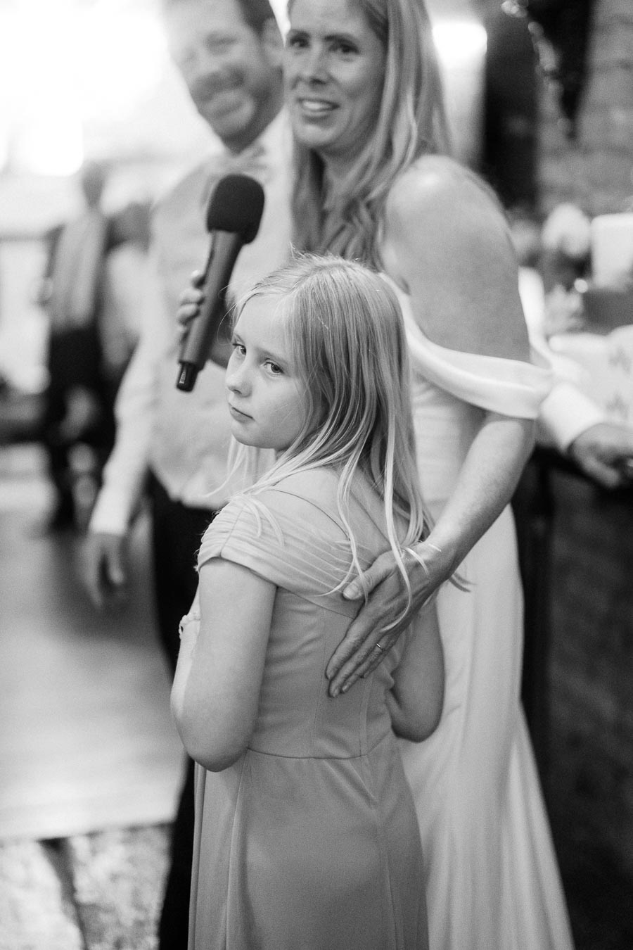 A black and white image capturing a heartfelt moment at a wedding reception. An elegantly dressed woman embraces a young girl next to a man holding a microphone. The setting appears intimate and warm, reflecting a special family gathering.