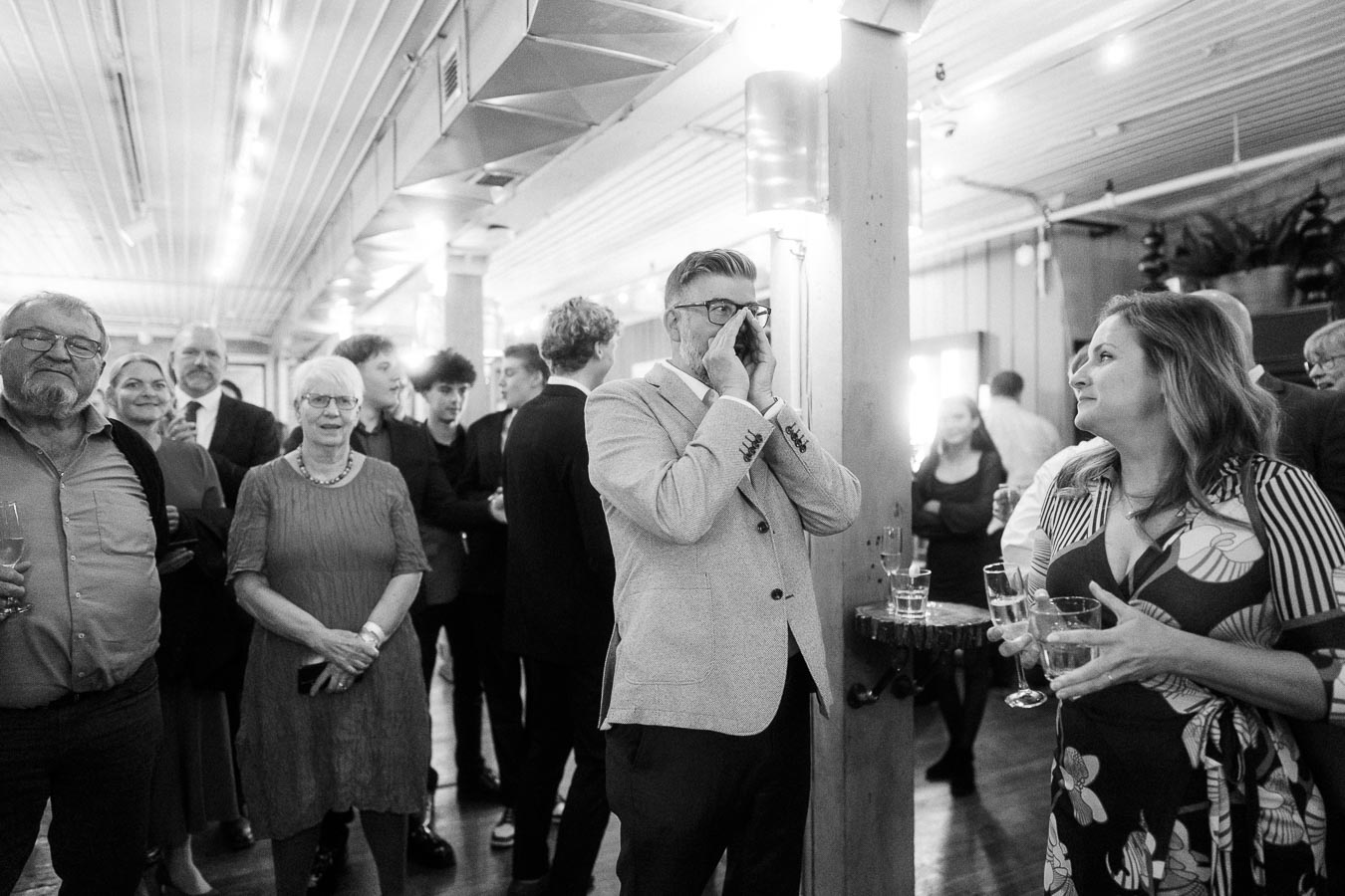 Black and white photo of a lively gathering at an indoor event; a man in a suit is speaking loudly, grabbing attention, while others in formal attire socialize and drink, evoking a celebratory atmosphere.