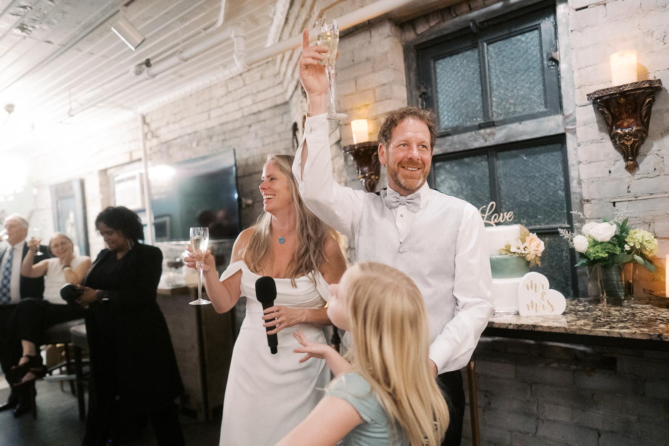 Happy couple raising champagne glasses in a celebratory toast at a wedding reception, surrounded by guests and a wedding cake with Mr. & Mrs. sign in a cozy, rustic setting.
