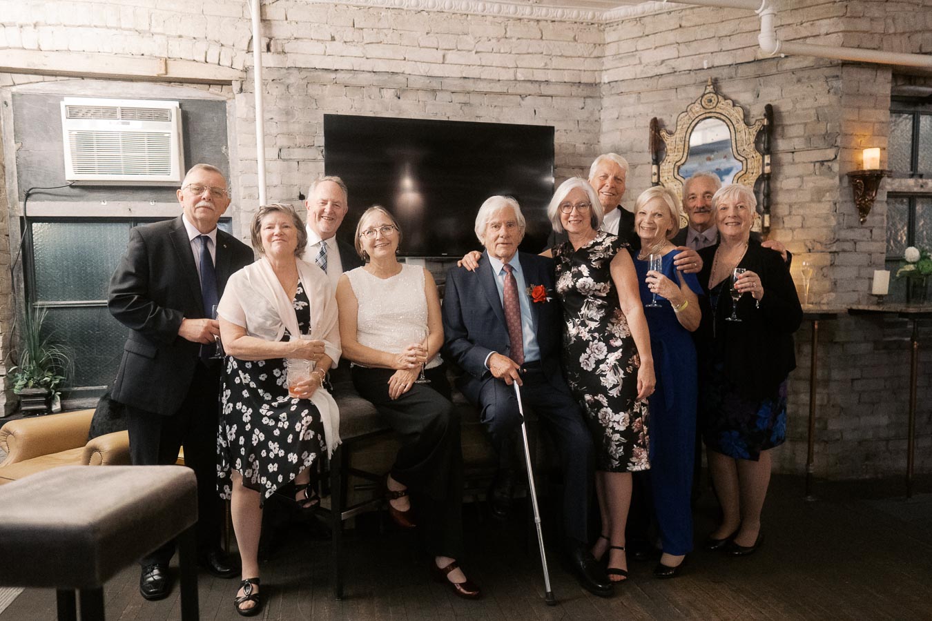 A group of people dressed elegantly poses for a photo in a warmly lit room with rustic brick walls, showcasing a festive gathering or celebration.