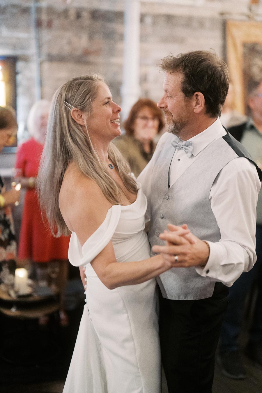 A happy couple dancing at a wedding reception, with the bride in a white gown and the groom in a gray vest and bowtie, surrounded by smiling guests in a warmly lit venue.