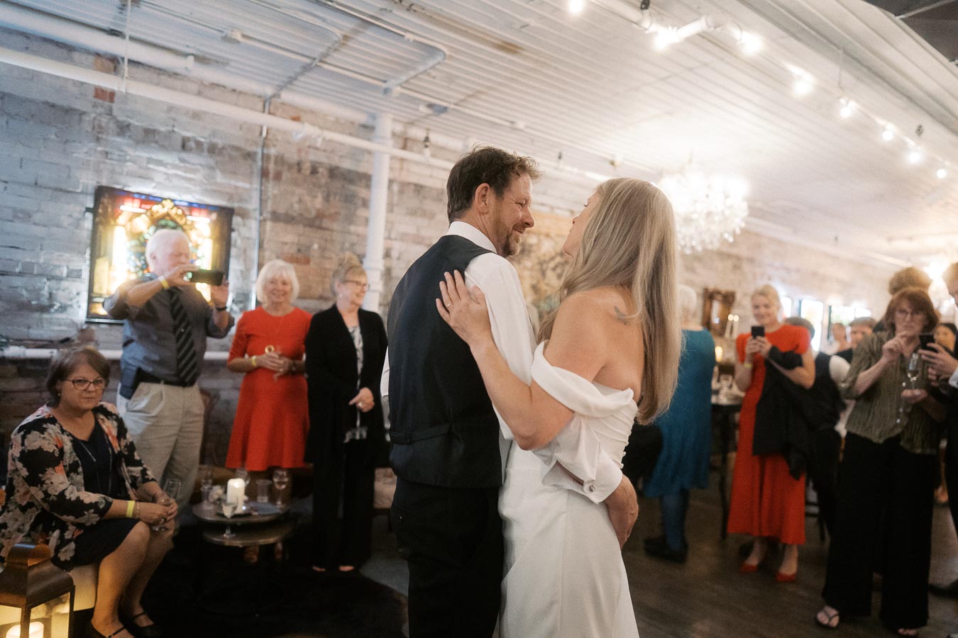 A couple dancing at a wedding reception with guests smiling and taking photos in a warmly lit, rustic venue.