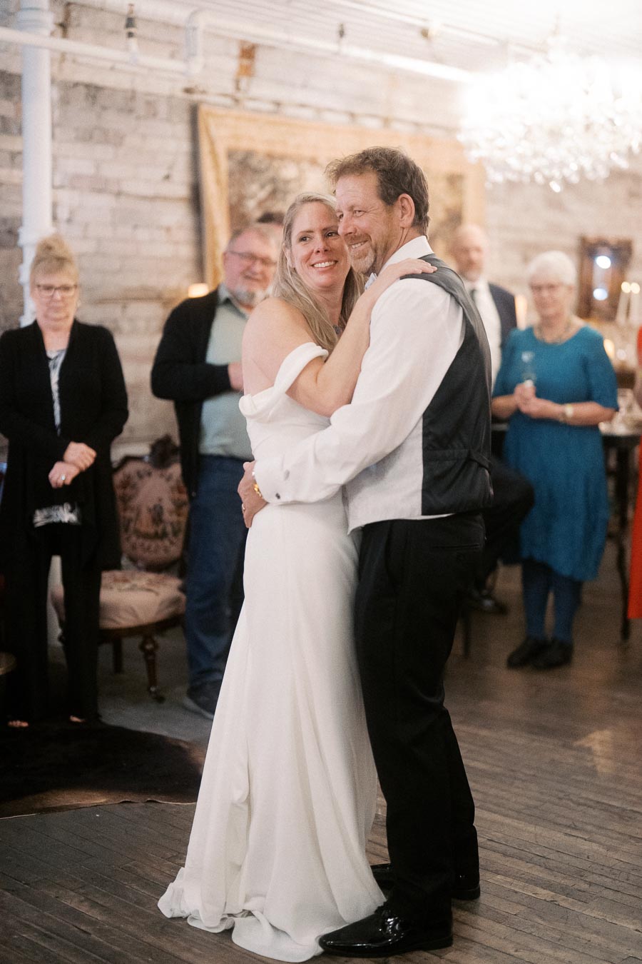 A joyful couple sharing a first dance at their wedding reception, surrounded by smiling guests in a warmly lit, rustic venue.