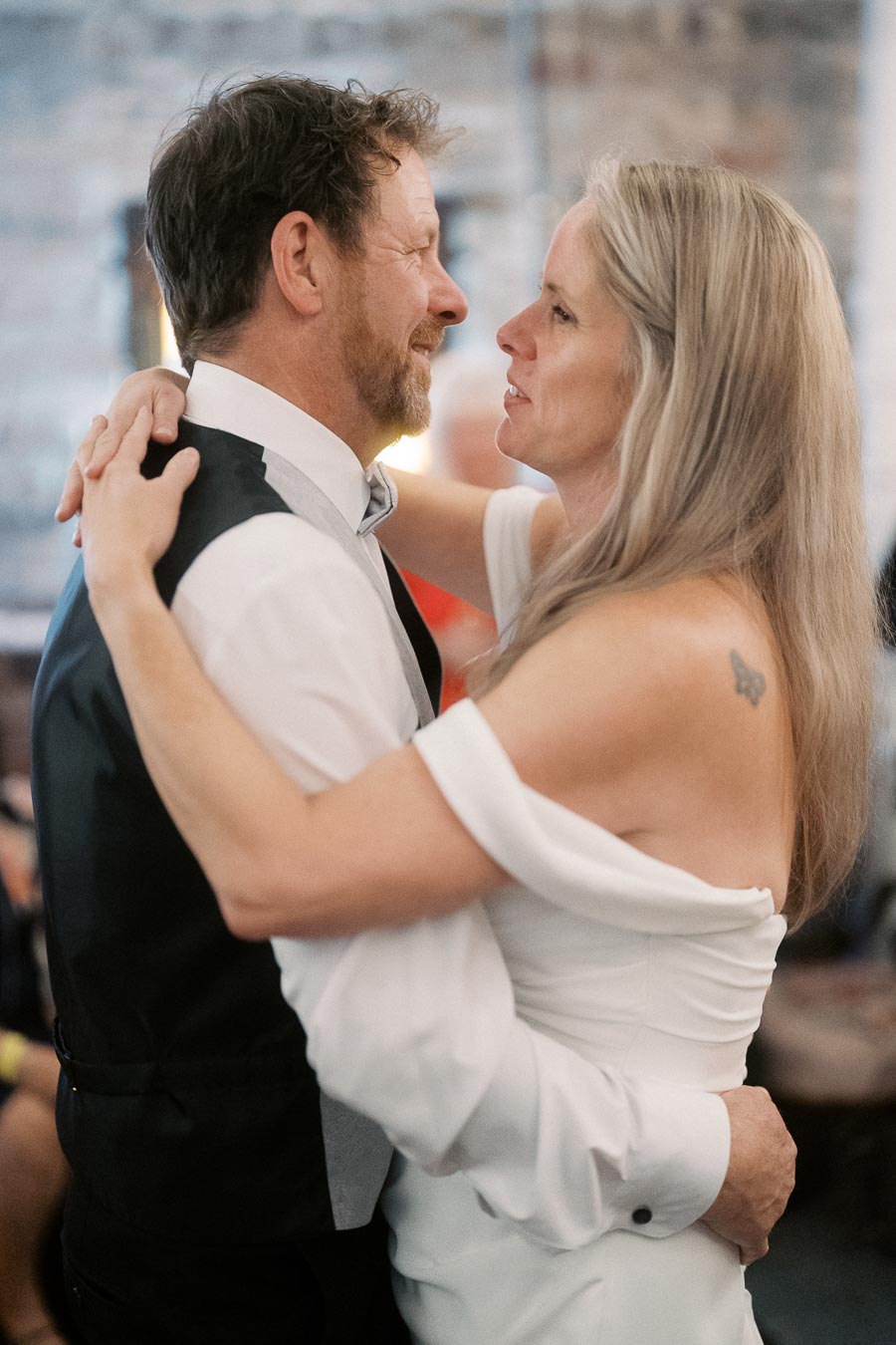 A couple shares a heartfelt dance at a wedding reception, with the woman in a white dress embracing the man in a formal vest and tie, creating a romantic and intimate moment.