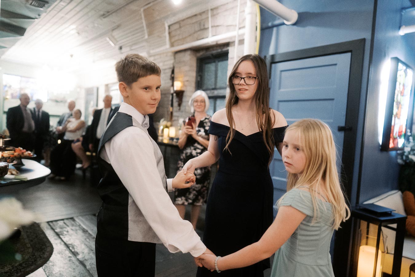 Three children holding hands and forming a circle during a family gathering in a warmly lit room.