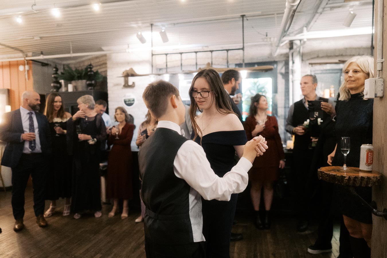 A couple dancing at an indoor social event, surrounded by elegantly dressed guests holding drinks, in a warmly lit, rustic venue.
