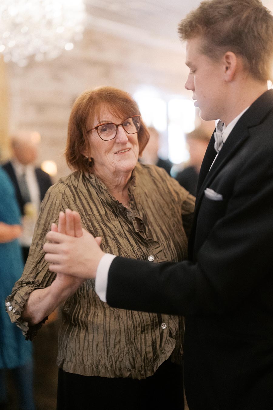 Elderly woman smiling while dancing with a younger man at a formal event, both dressed in elegant attire, with a blurred festive background.