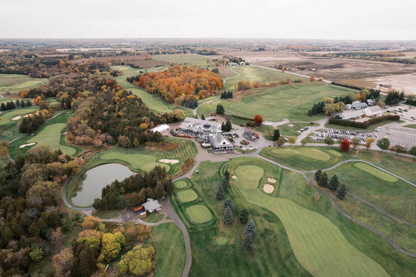 Aerial view of a picturesque golf course surrounded by lush greenery and autumn foliage, featuring a clubhouse, ponds, and sand bunkers, with expansive farmland visible in the background.
