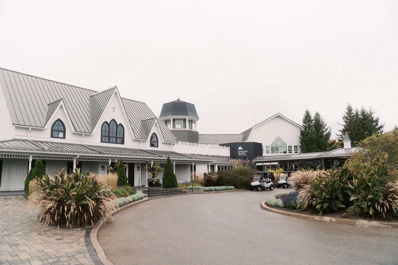 Exterior view of a modern golf clubhouse with a pitched roof and large windows, surrounded by landscaped gardens and golf carts parked at the entrance.