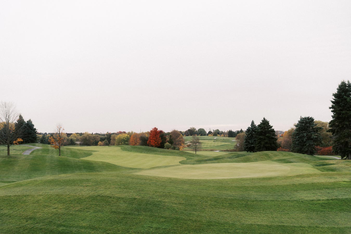 Scenic view of a lush green golf course surrounded by colorful autumn trees, featuring rolling fairways and a cloudy sky.