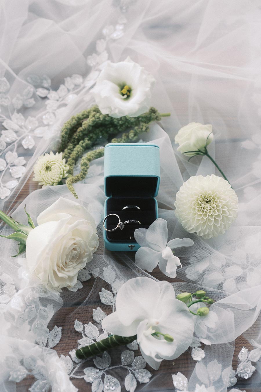 Elegant wedding rings in a turquoise box surrounded by white flowers and delicate lace fabric, creating a romantic and sophisticated atmosphere.