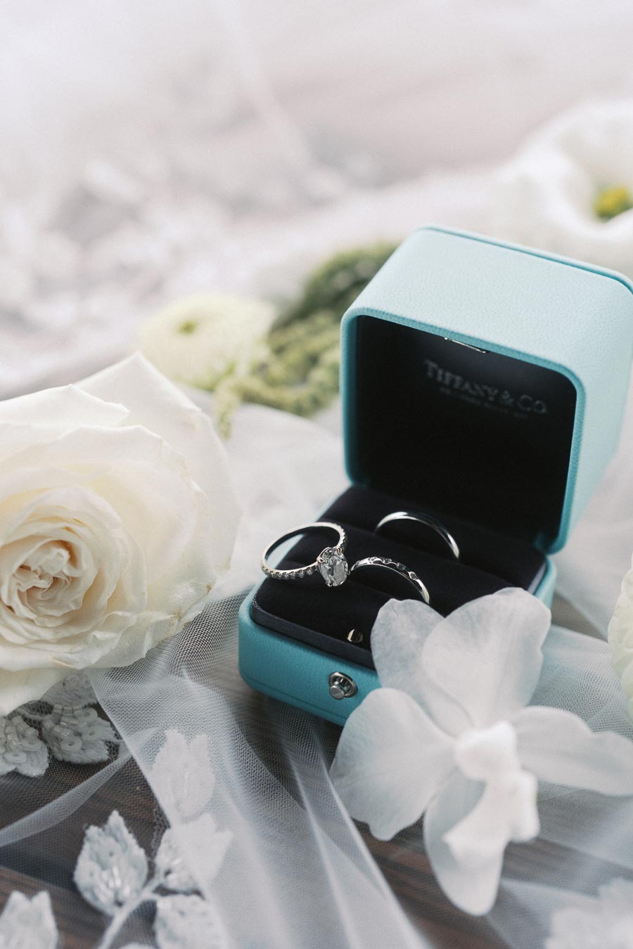 A silver engagement ring with a diamond setting displayed in a light blue jewelry box from Tiffany & Co., surrounded by delicate white roses and elegant lace fabric.