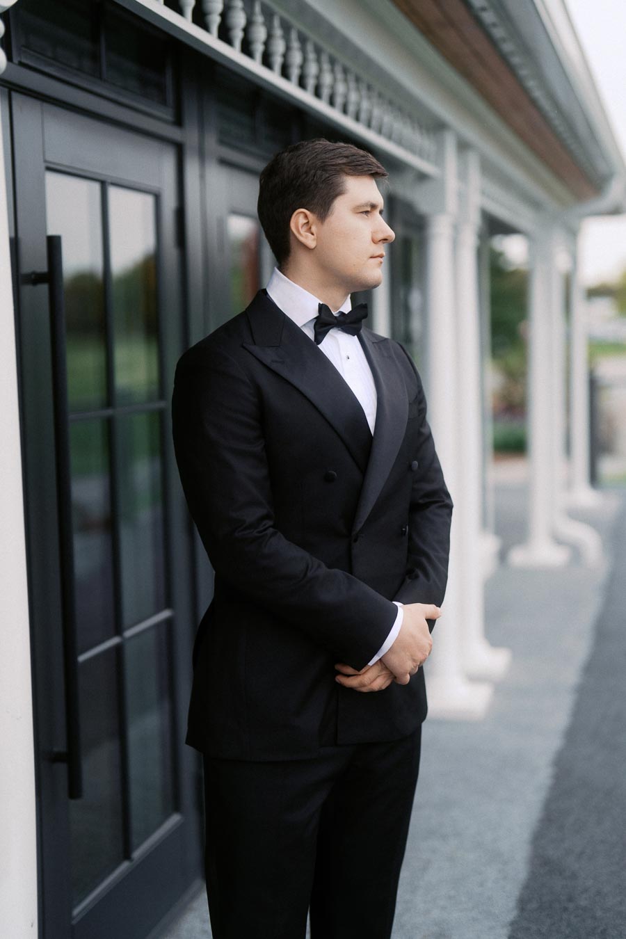 A man in a formal black tuxedo stands elegantly on a veranda, gazing into the distance, with architectural features like columns and large windows in the background.