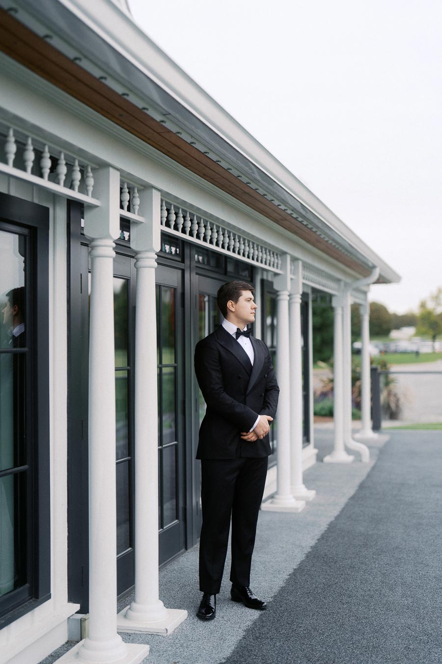 Elegant man in a black tuxedo standing on a stylish veranda with white columns, looking thoughtfully into the distance.