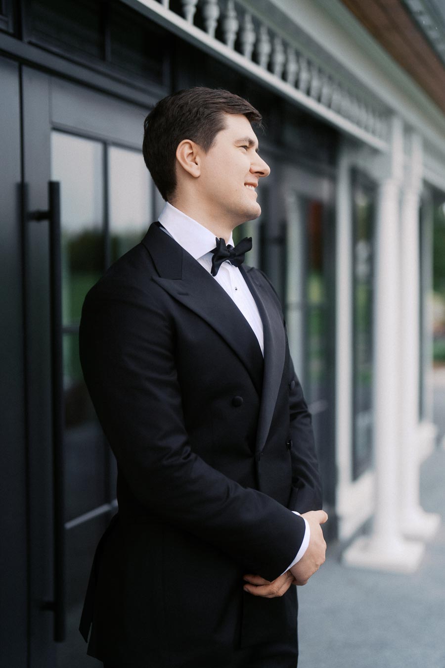 A man in a formal black tuxedo with a bow tie stands confidently outside a classic building with white columns.
