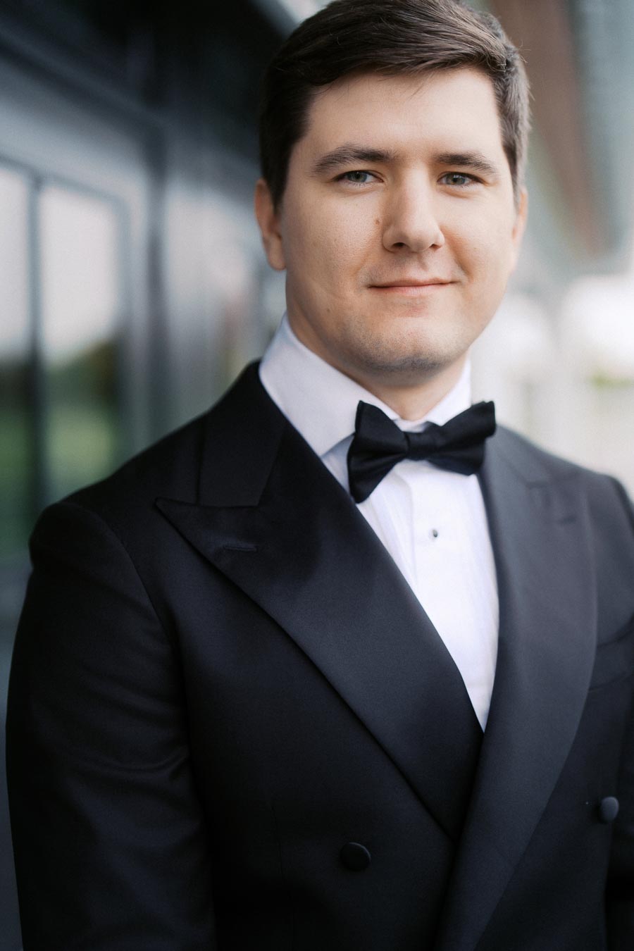 Portrait of a man in a formal black tuxedo with a bow tie, standing outdoors with a blurred background.