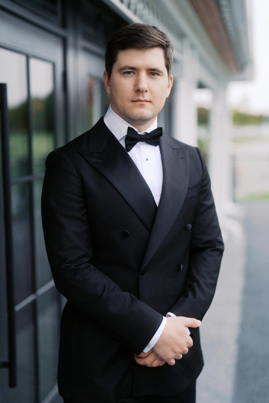 A man in a formal black tuxedo with a white dress shirt and black bow tie standing confidently outdoors near a modern building, exuding elegance and sophistication.