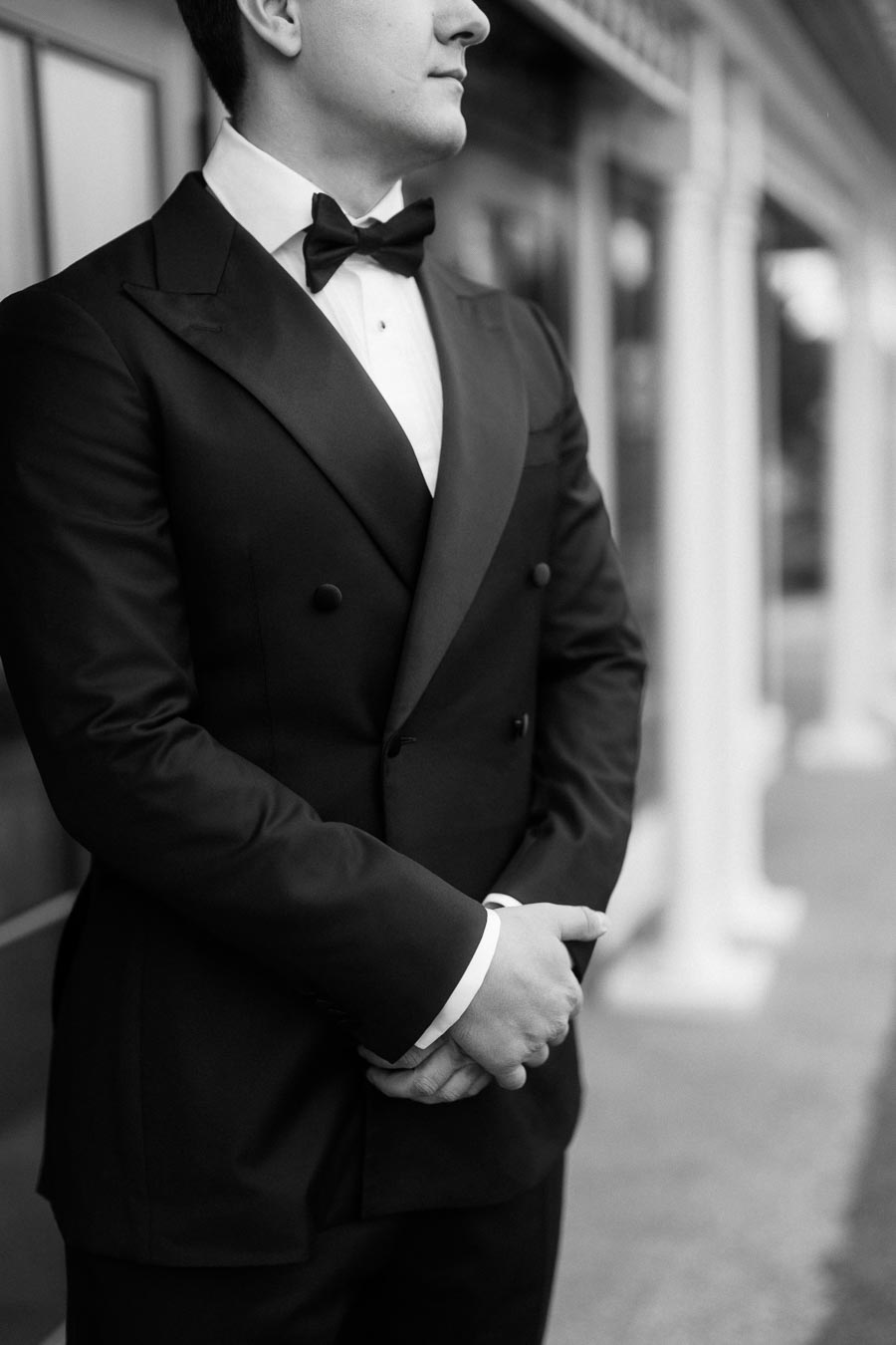 A well-dressed man in a formal black tuxedo with a bow tie stands confidently with hands clasped in front, showcasing elegance and style.
