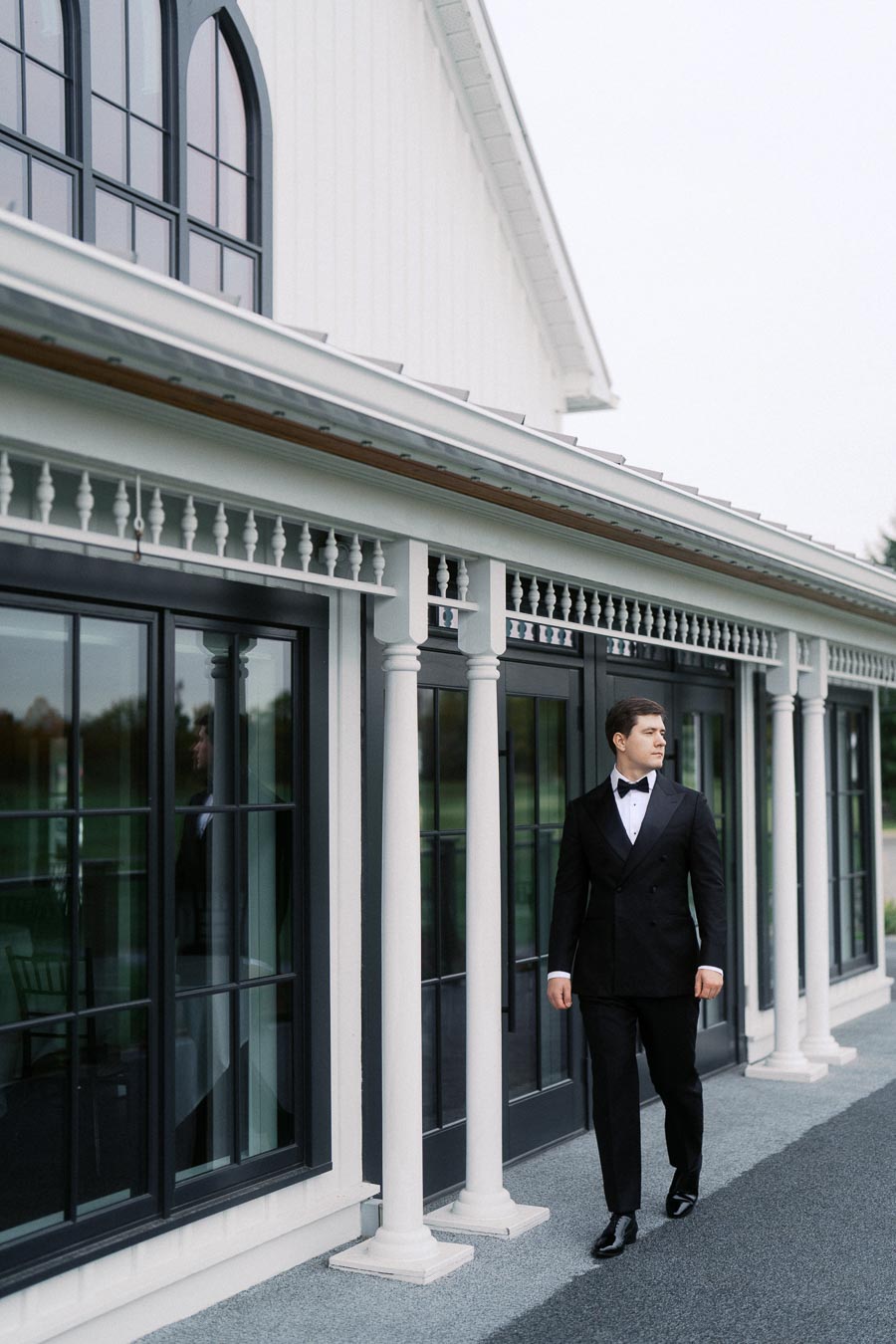 Man in a formal black tuxedo walking outside a stylish building with large windows and classic white columns.