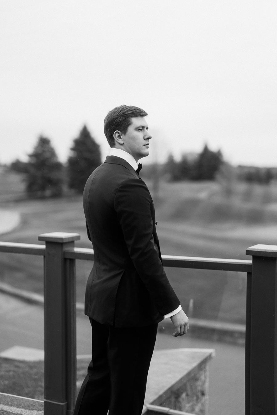 A person in formal attire, standing on a terrace overlooking a landscaped area, gazing into the distance, in a black and white photograph.