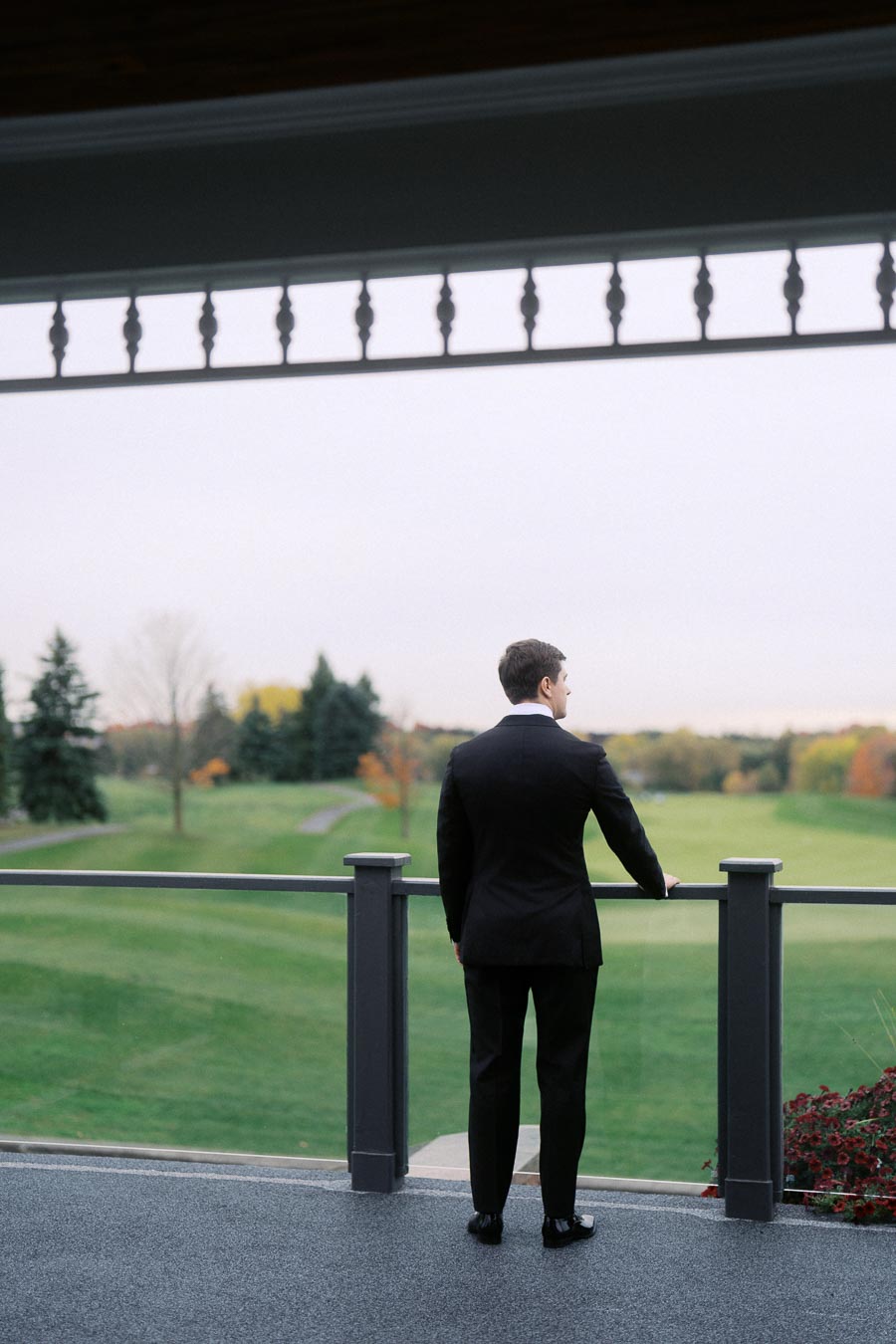 Man in a black suit standing on a balcony overlooking a lush green landscape with trees in the distance.