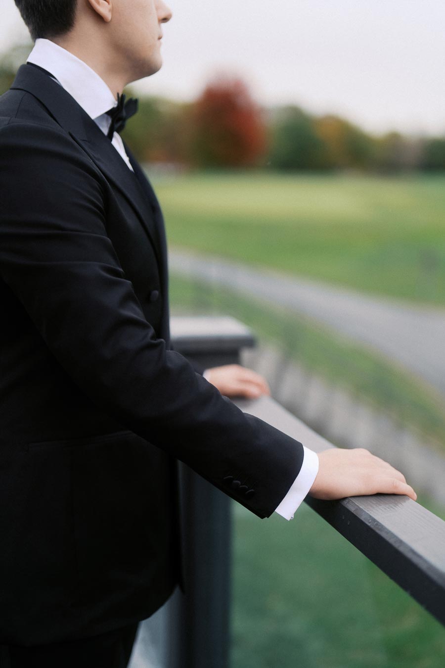 Man in a black tuxedo standing on a balcony overlooking a blurred green landscape with fall foliage in the background.